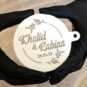 May include: A white, circular ornament with the names "Khalid & Rabiaa" and the date "25.11.22" etched in a decorative font. The design includes a circular border and leaf accents. The ornament is held by hands wearing black gloves.