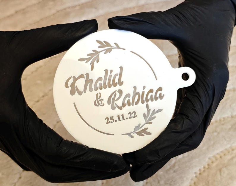 May include: A white, circular ornament with the names "Khalid & Rabiaa" and the date "25.11.22" etched in a decorative font. The design includes a circular border and leaf accents. The ornament is held by hands wearing black gloves.