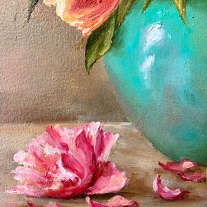 May include: A single pink peony flower with petals scattered around it on a light brown surface. The flower is in focus, while the background is blurred. A turquoise vase with a bouquet of orange tulips is visible in the background.