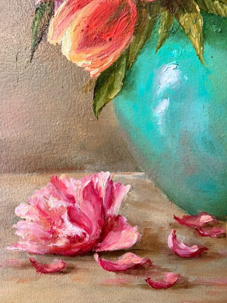 May include: A single pink peony flower with petals scattered around it on a light brown surface. The flower is in focus, while the background is blurred. A turquoise vase with a bouquet of orange tulips is visible in the background.