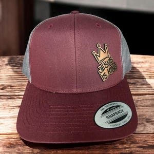 Puede incluir: Una gorra de béisbol de color granate y gris con una corona dorada y las palabras "Jesus is King" en la parte delantera. La gorra tiene una parte trasera de malla y una etiqueta "The Authentic Yupoong Snapback" en la parte posterior.