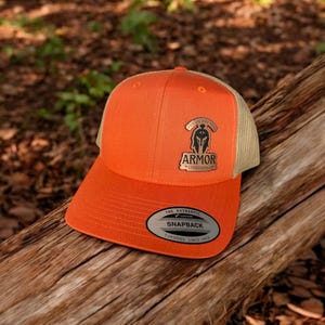 Peut inclure: Casquette de baseball orange et beige avec un écusson noir et or qui dit "Mettez l'armure complète de Dieu Éphésiens 6:11" et "Armure Éphésiens 6:11". La casquette a un autocollant noir et argent qui dit "The Authentic Snapback Yupoong Since 1974".