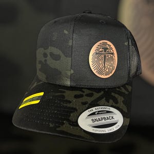 Peut inclure: Casquette de camionneur camouflage noir et vert avec un patch de couleur cuivre représentant un marteau et un clou. Le patch est circulaire et présente un motif d'empreinte digitale. La casquette a un dos en maille noire et une fermeture à pression noire. La casquette porte également une étiquette blanche avec le texte "THE AUTHENTIC YUPOONG SNAPBACK YUPOONG SINCE 1974".