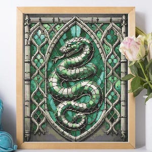 May include: A framed cross-stitch artwork featuring a green and white snake in a stained-glass style design. The artwork is set against a green background with ornate silver-colored detailing. The frame is made of light-colored wood.
