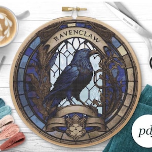 May include: A cross-stitch embroidery hoop featuring a blue raven in a stained-glass window design. The word "RAVENCLAW" is arched across the top. The design is set in a wooden hoop, with a PDF logo in the corner.