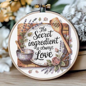 Puede incluir: Un bordado de punto de cruz en un aro de madera presenta el texto "The Secret Ingredient is always Love". El diseño incluye una cuchara de madera, un cuenco y elementos florales. La paleta de colores incluye tonos de marrón, morado y blanco.