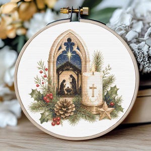 May include: A circular wooden embroidery hoop containing a cross-stitch pattern of a nativity scene inside a gothic archway. The design features a lit candle with a cross, a pinecone, holly leaves with red berries, and a star on a white fabric.