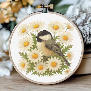 May include: Cross-stitch embroidery in a wooden hoop. The design features a small bird with black and gray feathers perched among white daisies with yellow centers and green leaves. The embroidery is set against a white background.