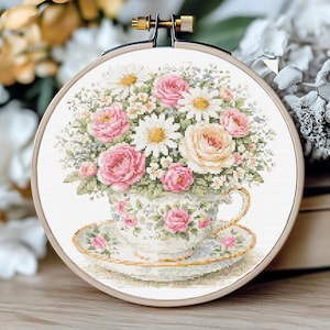 Puede incluir: Bordado de punto de cruz en un aro de madera. El diseño presenta un arreglo floral de rosas rosas, margaritas y otras flores en una taza y un platillo. La taza y el platillo son blancos con un borde dorado.