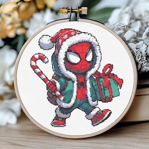 May include: A cross-stitch embroidery hoop featuring a cartoon Spider-Man character in a Santa hat and winter coat. He holds a candy cane and a wrapped gift. The design is in red, green, white, and black.