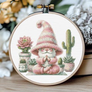 May include: A pink and white gnome meditating in a yoga pose surrounded by succulents and cacti. The gnome is wearing a pink hat and has a white beard.