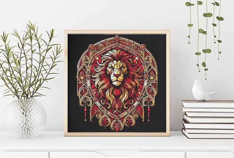 May include: Cross-stitch artwork in a wooden frame, depicting a lion's head with a red and gold mane on a black background. The design is framed in a light wood tone. The artwork is displayed on a white surface.
