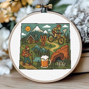 May include: A cross-stitch embroidery hoop featuring a pixelated outdoor scene. The design includes a hiker, a mountain biker, a cabin, mountains, a lake, and a beer mug. The color palette includes greens, browns, blues, and yellows, with a smiling sun in the sky.