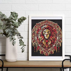 May include: A framed art print showing a detailed illustration of a lion's head. The lion has a red mane and gold accents, set against a black background. The artwork is framed in a white border and rests on a wooden table.