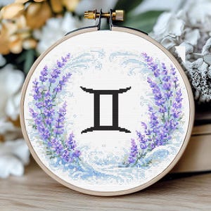 Peut inclure: Un cercle à broder avec le symbole du signe astrologique Gémeaux en noir. Le motif est entouré d'une couronne de fleurs de couleur lavande. Le fond présente un motif bleu clair en forme de vagues, le tout sur un tissu blanc.