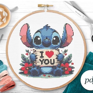 May include: Cross stitch pattern featuring a blue Stitch character holding a sign that says "I love you" with a red heart. The character is surrounded by green leaves and red flowers.