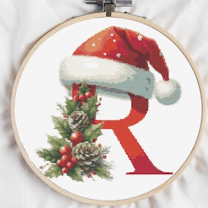 Puede incluir: Bordado de punto de cruz en un aro de madera con la letra "R" roja con un gorro de Papá Noel, acebo, piñas y bayas. El gorro tiene ribete blanco y un pompón blanco. El diseño está sobre un fondo blanco.