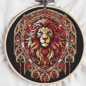 May include: A cross-stitch embroidery featuring a lion's head in a decorative frame. The lion's mane is red and yellow, with a detailed face. The frame is red and gold on a black background, set in a wooden embroidery hoop.