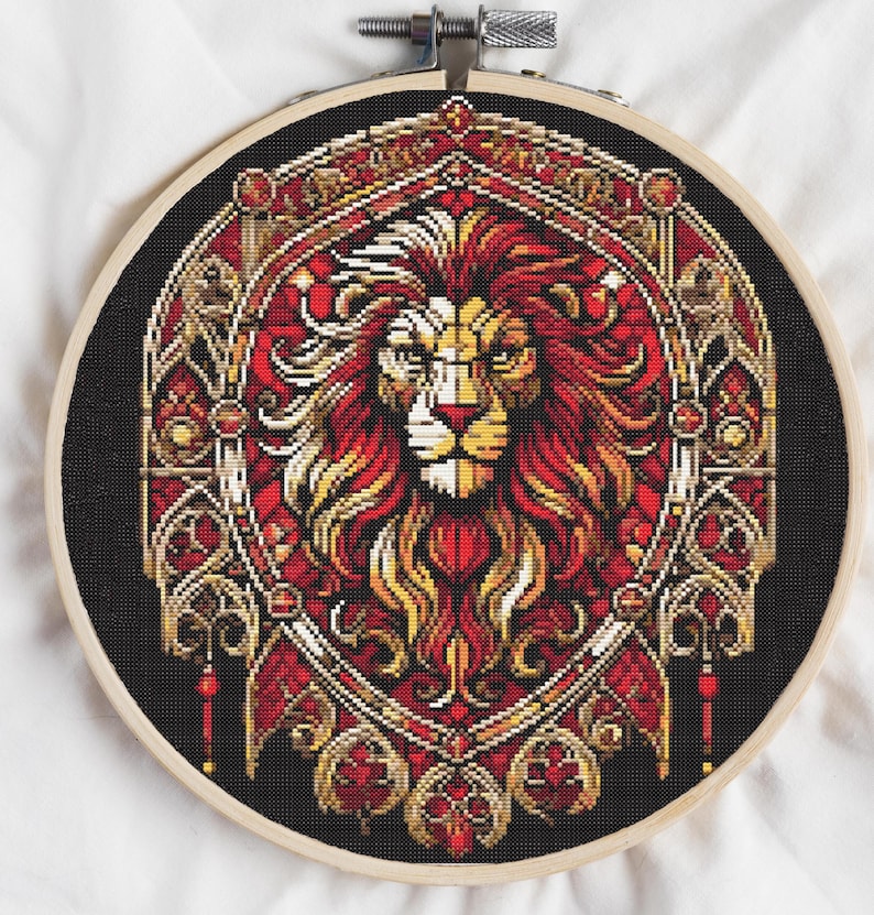 May include: A cross-stitch embroidery featuring a lion's head in a decorative frame. The lion's mane is red and yellow, with a detailed face. The frame is red and gold on a black background, set in a wooden embroidery hoop.