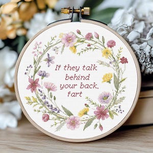 May include: A cross-stitch embroidery hoop with a floral wreath design. The text reads, "If they talk behind your back, fart." The wreath features pink, yellow, and purple flowers with green foliage. The hoop is made of wood and has a brass-coloured tightening mechanism.