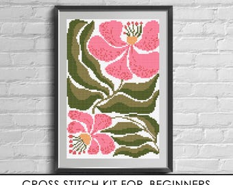 May include: A cross stitch kit for beginners featuring a printed pattern of two large pink flowers with yellow centers and green leaves, including canvas and threads. The design is displayed in a black frame against a white brick wall background.