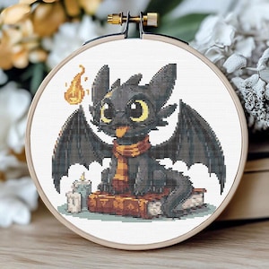 Puede incluir: Bordado de punto de cruz de un dragón negro con grandes ojos amarillos, alas y una bufanda, sentado sobre libros. Una pequeña llama y una vela también están incluidas en el diseño. El bordado está en un aro de madera.
