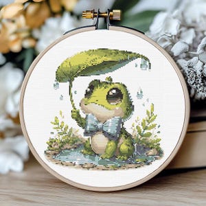 Op de afbeelding: Kruissteekborduurwerk van een kikker die schuilt onder een bladerenparaplu. De kikker is groen met een blauwe vlinderdas, omringd door groen en regendruppels. De borduursel zit in een houten borduurring.