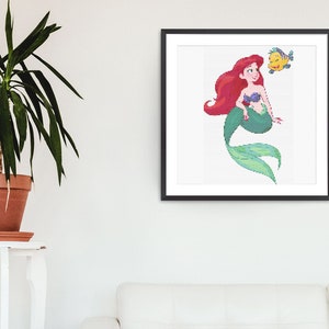 May include: A framed art print showing a pixelated illustration of a mermaid with red hair, a purple seashell top, and a green tail. A yellow fish is next to the mermaid. The artwork is displayed on a white wall.