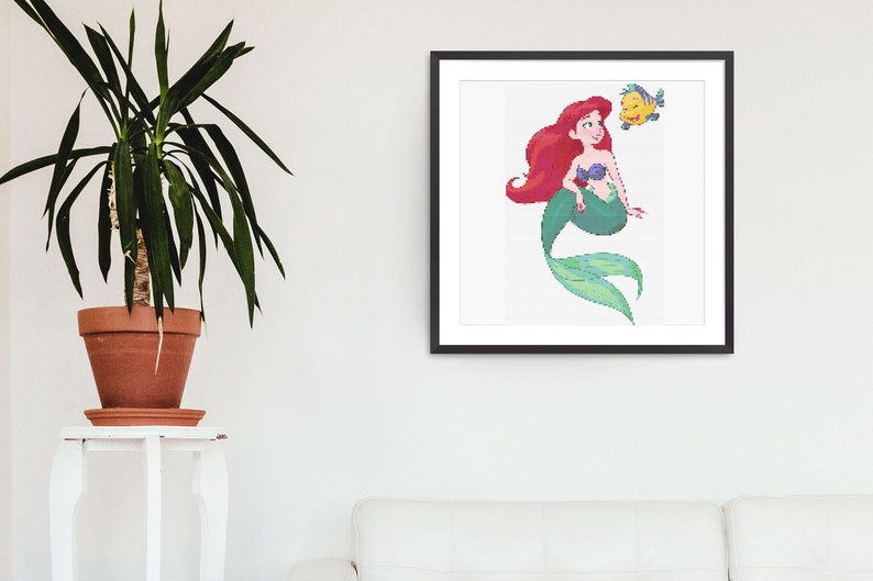May include: A framed art print showing a pixelated illustration of a mermaid with red hair, a purple seashell top, and a green tail. A yellow fish is next to the mermaid. The artwork is displayed on a white wall.