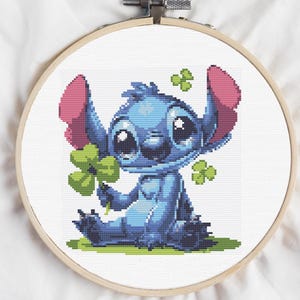 Puede incluir: Personaje azul Stitch sosteniendo un trébol de cuatro hojas con orejas rosas y una expresión feliz. El personaje está sentado en un parche de hierba verde con más tréboles alrededor.