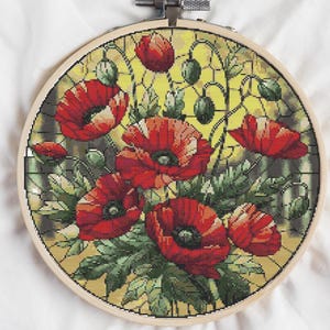 May include: A cross-stitch embroidery hoop with a colorful design of red poppies in a stained glass style. The background is a yellow and green pattern.