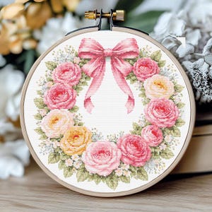 Op de afbeelding: Een borduurring met een bloemenkransontwerp. De borduurring toont roze en gele rozen, groene bladeren en een roze strik. De borduurring is geplaatst in een houten ring, een klassiek ambachtelijk item.