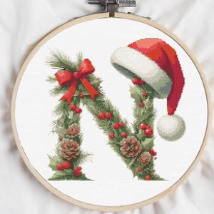 Puede incluir: Bordado de punto de cruz de la letra "N" decorada con vegetación navideña, bayas rojas, piñas y un lazo rojo. Un gorro de Papá Noel está colocado en la parte superior derecha. El bordado está en un aro de madera.