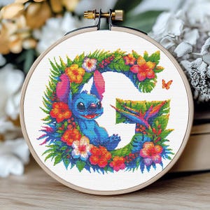 Könnte beinhalten: Kreuzstich-Stickerei in einem Holzreifen mit dem Buchstaben "G", umgeben von einem Blumenkranz. Eine blaue Cartoon-Figur ist in dem Buchstaben eingebettet, mit bunten Blumen und einem Schmetterling, der das Design akzentuiert. Die Stickerei befindet sich auf weißem Stoff.