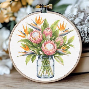 May include: A cross-stitch embroidery project in a circular wooden hoop featuring a bouquet of pink protea flowers and orange bird of paradise flowers arranged in a clear glass mason jar, all stitched onto white fabric with green foliage.