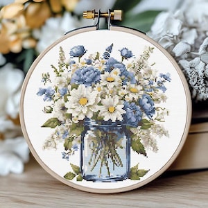 Può includere: Un telaio da ricamo a punto croce con una composizione floreale in un barattolo blu. Il bouquet comprende margherite bianche, rose blu e verde. Il ricamo è ambientato in un telaio di legno, creando un'estetica rustica.