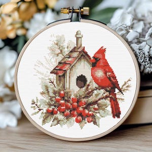 May include: A completed cross-stitch embroidery in a wooden hoop. The design features a red cardinal perched near a birdhouse with a red-tiled roof, surrounded by greenery and red berries. The background is white.