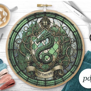 May include: A green and gold cross-stitch pattern of the Slytherin house crest from the Harry Potter series. The crest features a green snake with a crown on its head, surrounded by a green and gold shield with a diamond pattern. The words "Slytherin" are written in a banner below the crest.