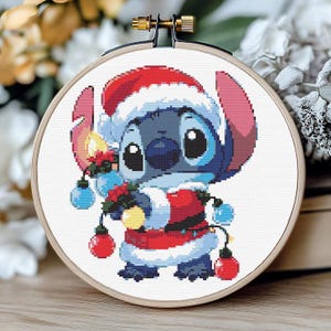 Op de afbeelding: Een kruissteekborduurwerk van Stitch uit Lilo & Stitch, gekleed in een kerstmanoutfit. De blauwe alien draagt een rode kerstmuts en jas, met kerstverlichting. De borduursel zit in een houten hoepel, ongeveer 15 cm in diameter.