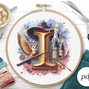 Puede incluir: Bastidor de bordado de punto de cruz con temática de mago. El diseño presenta la letra "I" dorada, un sombrero de mago, un búho, una varita mágica, velas y un castillo. La paleta de colores incluye dorado, azul y rosa.