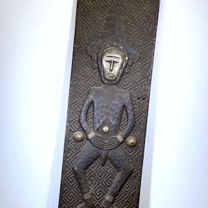 May include: A dark brown, rectangular wooden carving featuring a stylized human figure with a large hat and geometric patterns. The figure has a silver face and is adorned with metal accents. The carving is likely a decorative art piece.