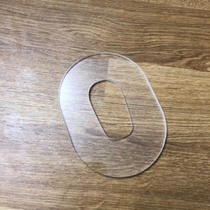 May include: A clear plastic stencil in the shape of the number zero. The stencil is on a wood surface.