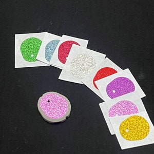 May include: Assortment of eight glitter-covered circles on white squares, with a single pink glitter circle on a grey background. The circles are in various colours, including green, blue, red, silver, purple, and gold.