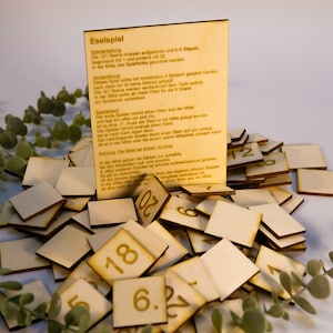 Peut inclure: Jeu de société en bois avec des tuiles carrées numérotées de 1 à 20, dispersées autour d'un panneau en bois avec du texte allemand. Le panneau contient les instructions du jeu. Des feuilles vertes sont en arrière-plan.
