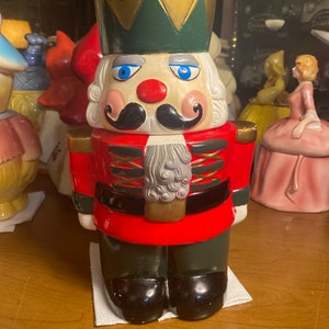 May include: A ceramic nutcracker cookie jar with a red and green uniform, a gold crown, and a white face with a black mustache. The nutcracker is standing on a white surface.
