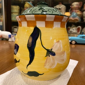 May include: A yellow ceramic cookie jar with a floral design. The jar has a green and white checkered rim and a green lid with a decorative pattern. The jar is sitting on a white paper towel.