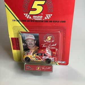 May include: A red, white, and yellow 1/64 scale die-cast stock car with a Kellogg's sponsorship. The car is in a plastic display case with a collector card featuring Terry Labonte, the 1996 NASCAR Champion. The card has a red, white, and blue background with the number 5 and the Hendrick Motorsports logo.