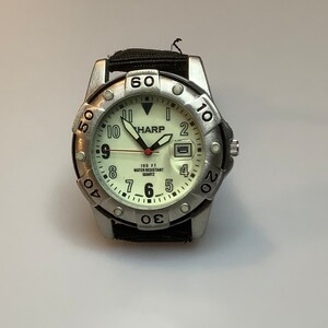 May include: A silver-toned SHARP quartz watch with a black fabric band. The watch face is off-white with black numerals and hands. The watch has a date display and is water-resistant.