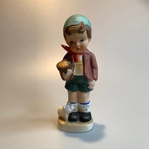 May include: A ceramic figurine of a young boy in a red jacket, green shorts, and a blue cap, holding a basket. The figurine has a detailed painted face and is standing on a small base with a white dog. A vintage collectible.