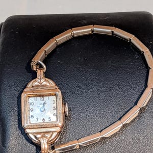 May include: A vintage gold-tone wristwatch with a square face, featuring a white dial with black Arabic numerals and the brand name Olev. The watch has a flexible, expandable metal link bracelet and a small winding crown on the side.